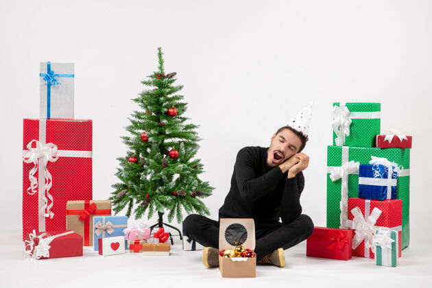 人年轻人坐在白色的墙上打哈欠度假礼物的正面图庆祝圣诞新郎