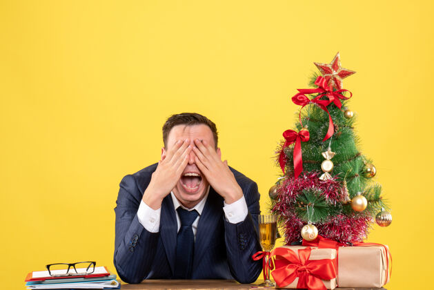 圣诞年轻人张着嘴捂着眼睛坐在圣诞树旁的桌子旁 用黄色的礼物男性开口树