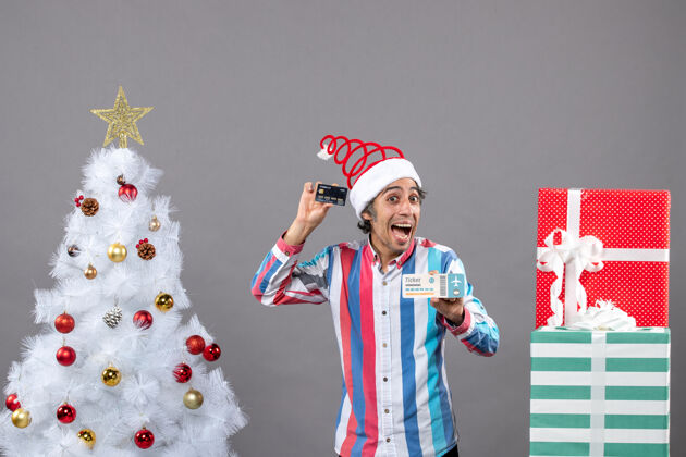 人正面图喜出望外的男子拿着卡片和旅行票围着圣诞树和礼物欣喜若狂肖像抱着