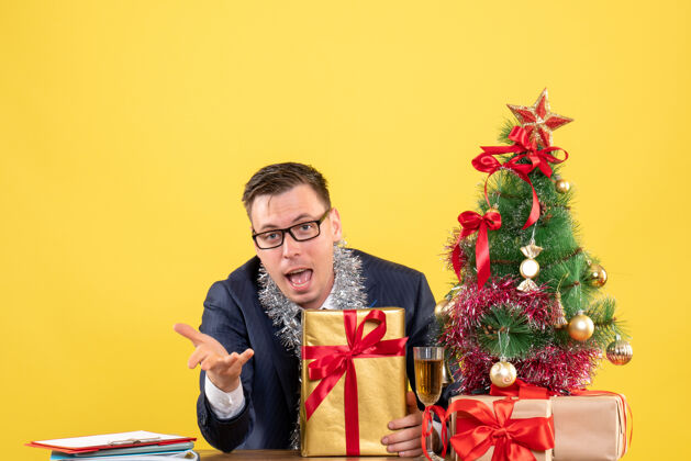 礼物拿着礼品盒的年轻人坐在圣诞树旁的桌子旁 拿着黄色的礼物拿着坐着人