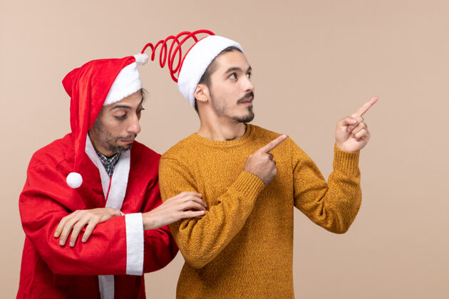 微笑正面图两个戴着圣诞帽的朋友一个挽着另一个的胳膊 另一个在米色孤立的背景上显示方向成人朋友帽子