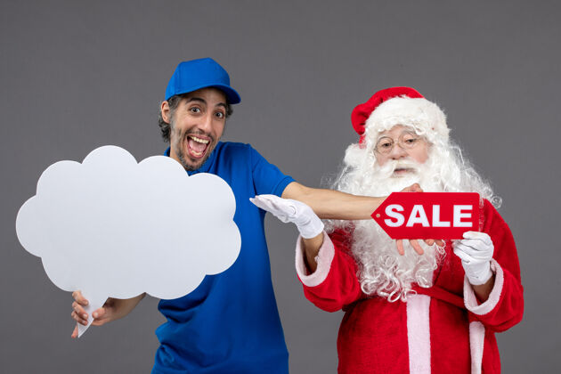 人圣诞老人的正面图 男信使手持云牌和灰色墙上的销售横幅前面帽子举行