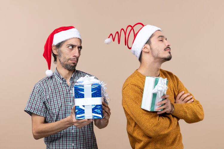 礼物正面图两个被冒犯的朋友戴着圣诞帽 拿着圣诞礼物 背景是米色的帽子风景男