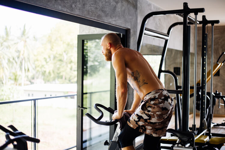 动力纹身 肌肉发达 胡须浓密的男人骑着自行车在体育馆里锻炼有氧运动 靠近大窗户 外面可以看到树运动机器健康