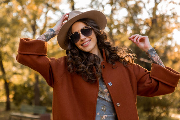 夹克迷人的时尚微笑的女人卷发漫步在街头公园穿着温暖的棕色外套秋季流行时尚 街头风格戴帽子和太阳镜配饰户外女性