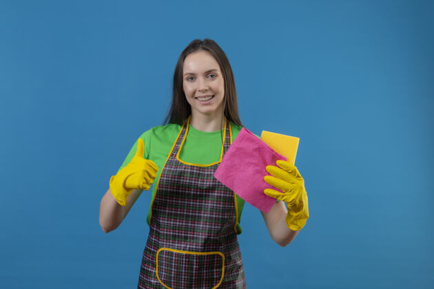 蓝色微笑着清洗穿着制服戴着手套拿着清洁工具的年轻女孩在孤立的蓝色背景上竖起大拇指年轻拇指工具