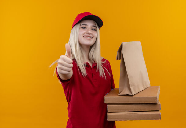 年轻微笑着送货的年轻女孩 穿着红色t恤 戴着帽子 戴着牙套 手里拿着披萨盒和纸口袋 大拇指竖立在孤立的橙色背景上口袋披萨帽子