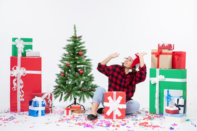 雪年轻女子围坐在礼物和白色墙上的小圣诞树前的视图情感装饰盒子