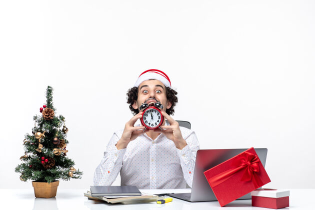 时钟戴着圣诞老人帽 拿着闹钟的年轻商人 正坐在白色背景的办公室里仔细地看东西帽子年轻商务人士
