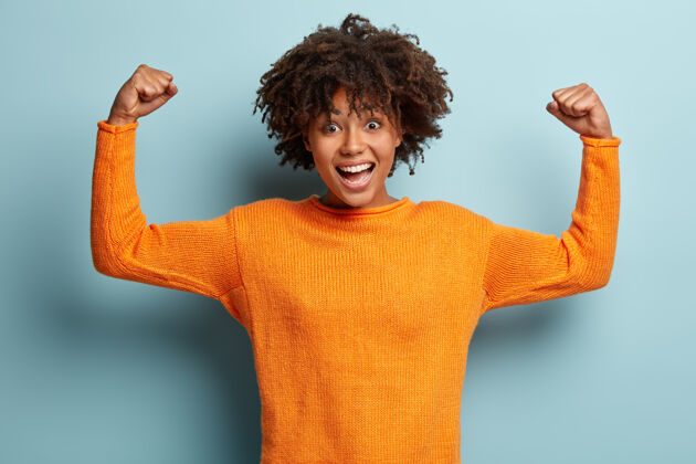 高兴快乐的美国黑人妇女卷发 举手 展示肌肉 展示她的力量 穿橙色套头衫肌肉成功美国