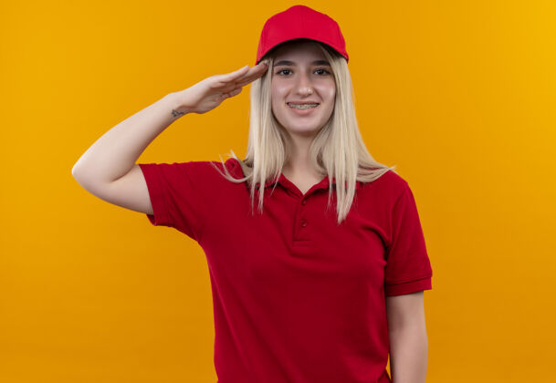 送货微笑的小女孩穿着红色t恤 戴着帽子 戴着牙套 在孤立的橙色背景上展示着敬礼的手势展示敬礼T恤