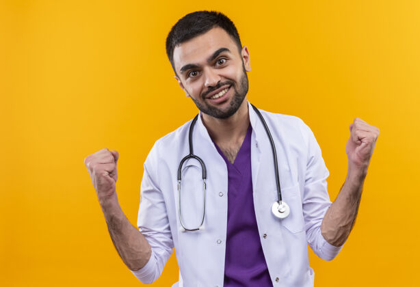 男性微笑的年轻男医生穿着听诊器医用长袍 在孤立的黄色背景上显示“是”的手势穿着礼服医学