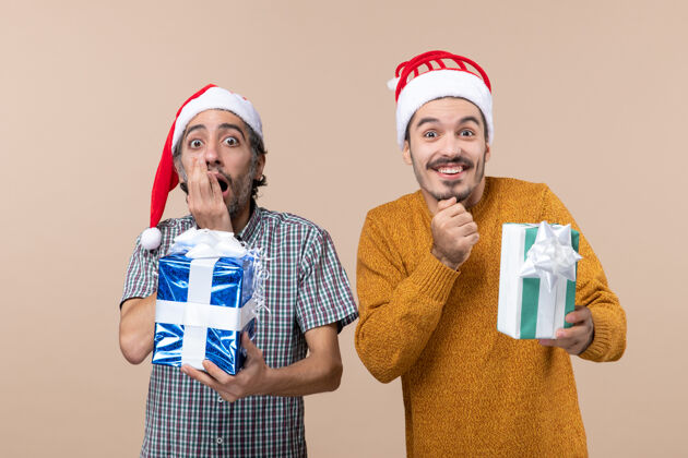 帽子正面图两个男人一个震惊 一个高兴地戴着圣诞帽 拿着他们的礼物在米色孤立的背景伙计们成人肖像