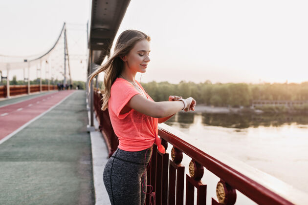 跑步穿着灰色裤子的苗条女孩站在户外看着智能手表欢快的欧洲女士戴着健身手镯在街上摆姿势有氧运动马拉松休息