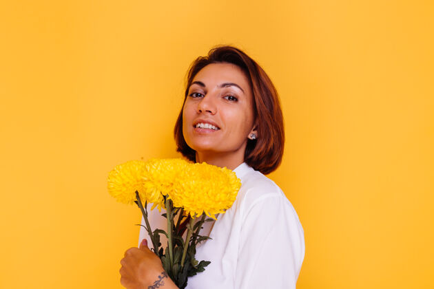 华丽摄影棚拍摄的黄色背景快乐的白人妇女短发穿着休闲服白衬衫和牛仔裤手持一束黄色紫苑植物礼物积极