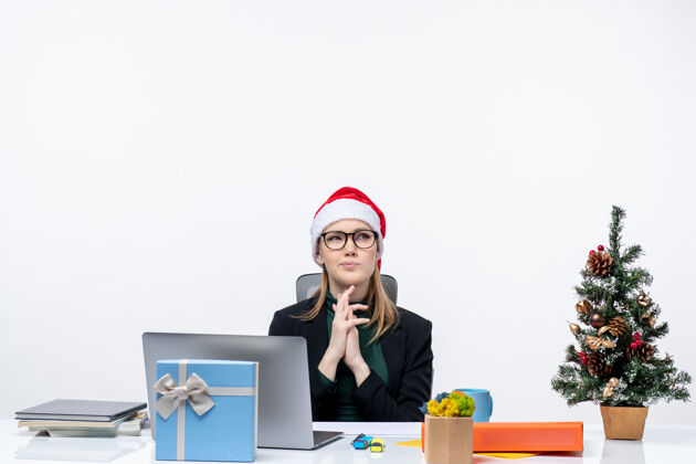 圣诞老人在白色背景的办公室里 一位戴着圣诞老人帽子的商务女士坐在一张桌子旁 桌子上放着圣诞树和礼物桌子女士帽子