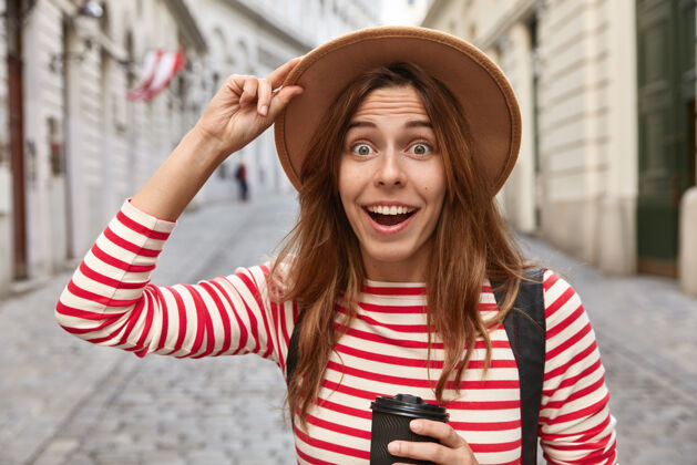 射击一个惊喜的欧洲女人手戴帽子 喝外卖咖啡 走在城市街道上的照片面部水平外卖