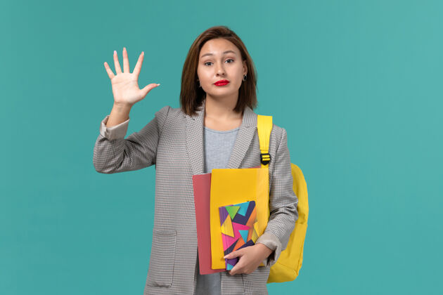 学生身穿灰色夹克 背着黄色背包 在浅蓝色墙上拿着文件和抄写本的女学生的正视图学校背包人