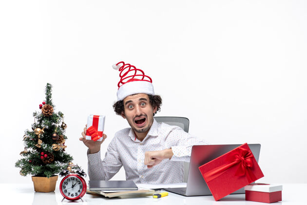 人圣诞气氛与紧张的年轻商人与圣诞老人帽子举行他的礼物 并检查他的白色背景时间抱着年轻礼物