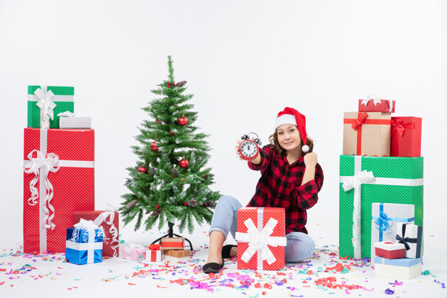 十二月前视图的年轻女子坐在圣诞礼物周围拿着白色墙上的时钟节日庆祝冬青