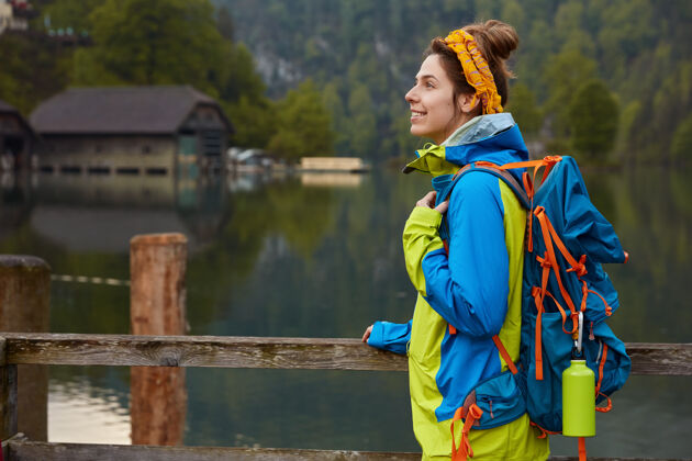 休闲开朗的女游客侧身镜头与小房子隔江傍湖的木桥相望 呼吸新鲜空气 享受大自然徒步旅行者背包森林