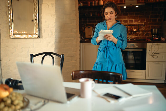 自由职业者身着蓝色连衣裙的严肃年轻女子站在厨房里拿着笔记本的室内肖像写作马克杯电子邮件