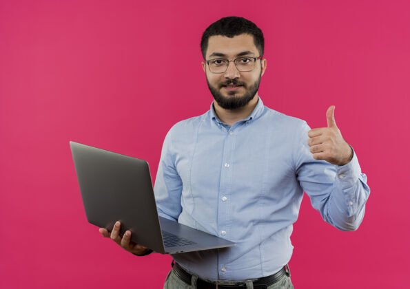 站着戴眼镜 穿蓝衬衫 留着胡子的年轻人拿着笔记本电脑微笑着竖起大拇指拿着年轻眼镜
