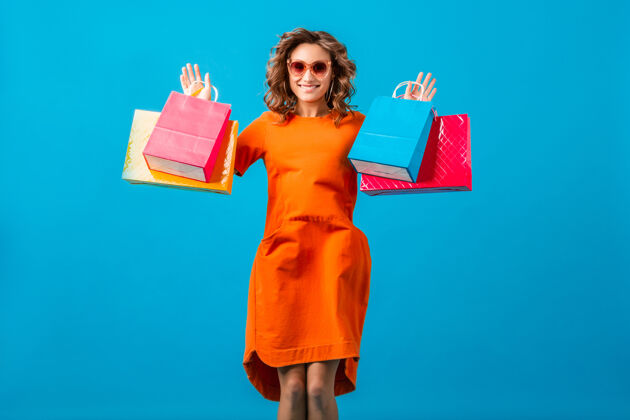 女性迷人的快乐情绪微笑时尚的女人购物狂在橙色时尚的超大礼服举行购物袋蓝色工作室背景隔离微笑太阳镜女士