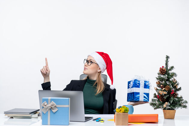 坐着圣诞节的心情与自信的年轻女子戴着圣诞老人的帽子 戴着眼镜坐在一张桌子上显示礼物指着上面的白色背景笔记本电脑指着秘书
