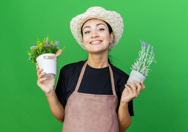 绿色年轻漂亮的女园丁围着围裙 戴着帽子 手里拿着盆栽植物 脸上洋溢着幸福的笑容抱着脸站着