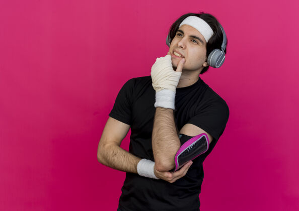 智能手机年轻的运动型男人 戴着运动服和头带 戴着手机和智能手机袖标 手放在下巴上 一边看着一边思考站着男人运动装