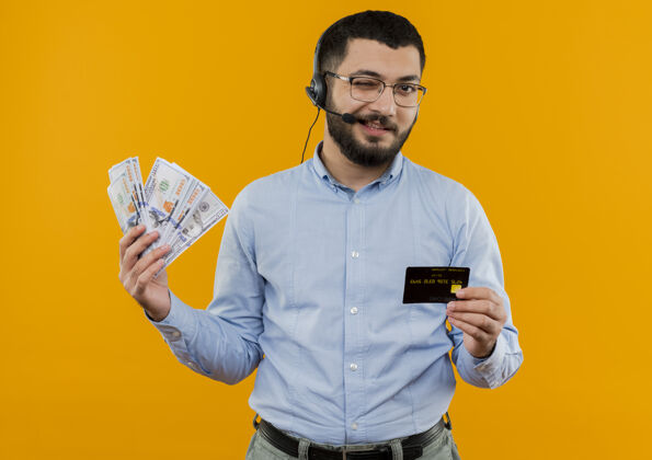 现金一个留着胡子的年轻人 穿着蓝色衬衫 戴着耳机 拿着麦克风 手里拿着现金 脸上露出信用卡的微笑和眨眼衬衫胡须显示