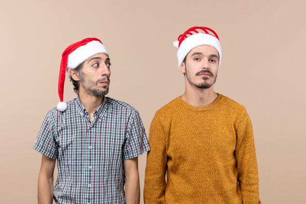 看前视图两个戴着圣诞帽的困惑的家伙一个看着另一个在米色孤立的背景上两个困惑的家伙帽子圣诞老人