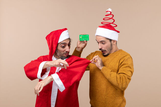 帽子前视图两个家伙一个穿着圣诞老人的外套 另一个拉着他的外套在米色孤立的背景上成人帽子外套