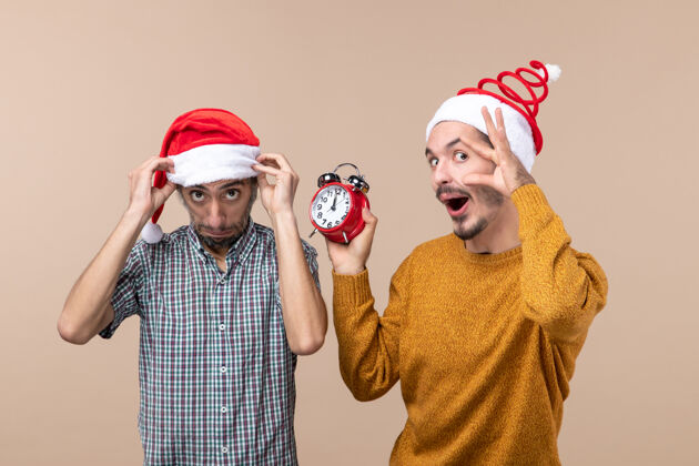 圣诞老人前视图两个男人一个拿着他的圣诞帽 另一个拿着闹钟 同时睁着眼睛在米色的孤立背景上打开拿着男人