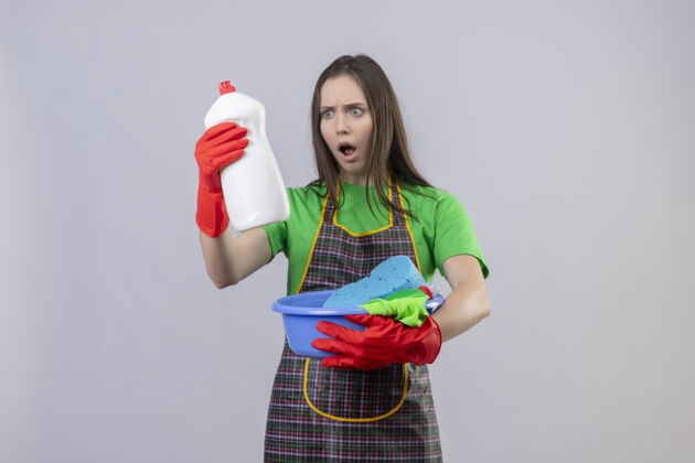 年轻害怕清洁的年轻女孩穿着制服 戴着红色手套 拿着清洁工具 看着她手上的清洁剂上孤立的白色背景女孩穿红