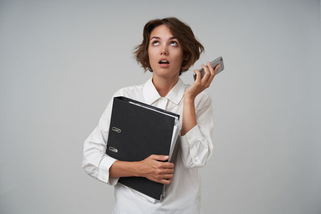 女性室内拍摄的是一位穿着正式服装 戴着智能手机和文件夹 拿着文件 谈吐无聊 眼睛打转的年轻黑发女子手机棕色智能手机