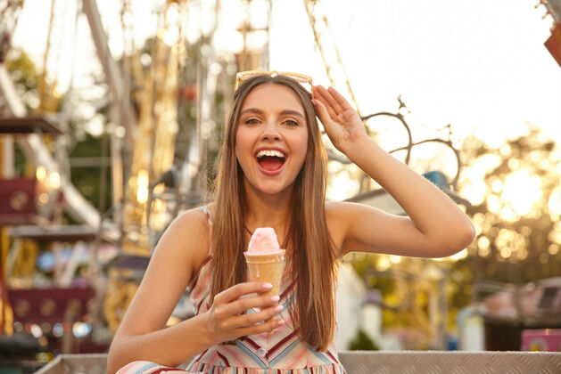 圆锥形年轻开朗的长发美女 头戴墨镜 面带笑容 一边吃着冰激凌 一边穿过公园里的景点 穿着夏装阳光年轻20多岁
