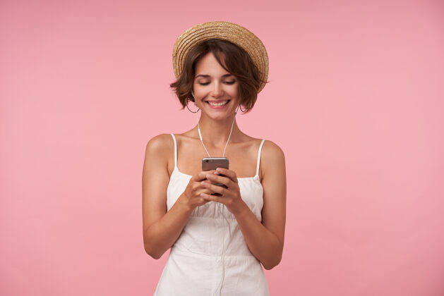 粉色开朗可爱的黑发女性 随意的发型看着手机屏幕 面带微笑 一边用耳机听音乐一边输入信息 与世隔绝肖像积极成人