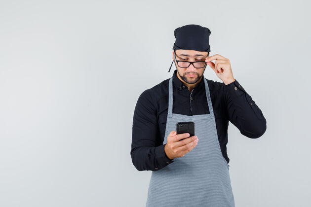 食物男厨师透过眼镜看着手机 穿着衬衫 围裙 看上去很忙前视图准备厨房职业