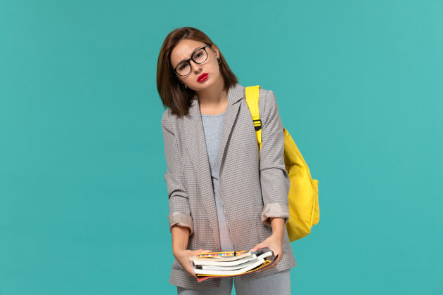 持有身着灰色夹克 黄色背包 蓝色墙壁上拿着书的女学生正面图Female大学成人