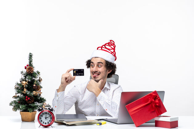 商务在白色背景的办公室里 戴着圣诞老人帽 拿着银行卡 伸出舌头的年轻忙碌的商人成人圣诞节持有
