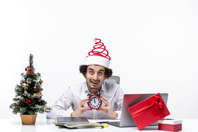 圣诞节年轻的留着胡须的滑稽商人戴着圣诞老人的帽子 拿着一个闹钟 惊讶地看着什么东西 坐在白色背景的办公室里男性办公室圣诞老人