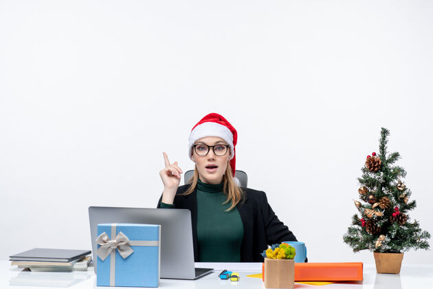 电脑一位带着圣诞老人帽子的商务女士坐在一张桌子旁 桌子上有一棵圣诞树和一份白色背景的礼物圣诞老人圣诞树体贴