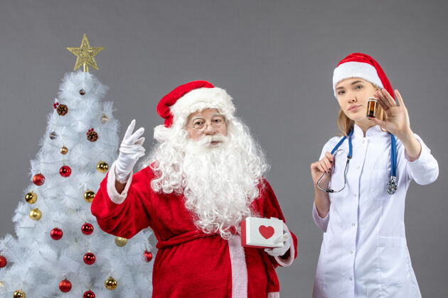 圣诞圣诞老人和女医生的前视图 女医生拿着装着药片的小瓶子在灰色的墙上圣诞老人圣诞老人服装