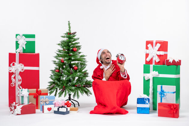 圣诞老人情绪激动的快乐圣诞老人坐在地上看上面 在礼物和白色背景装饰圣诞树附近展示时钟装饰礼物礼物