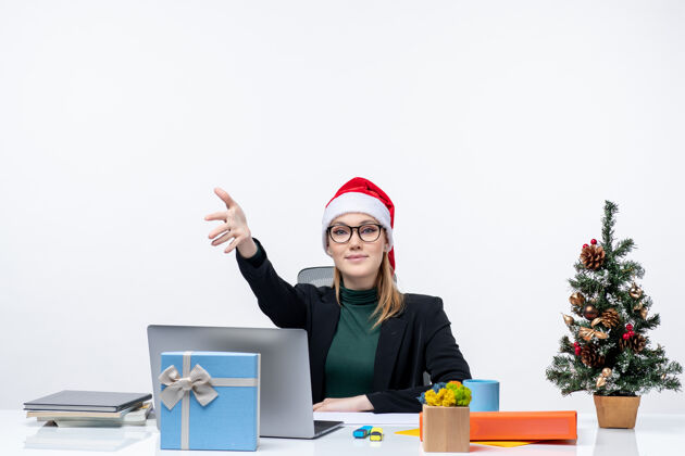 女人新年气氛：年轻漂亮的女士戴着圣诞老人帽坐在桌子旁 桌上放着圣诞树和礼物 欢迎办公室里的人圣诞树某人礼物