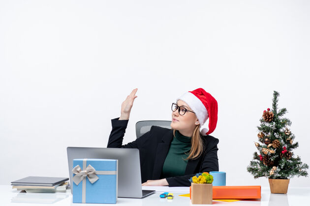 说新年气氛：年轻漂亮的女士戴着圣诞帽坐在桌子旁 桌上放着圣诞树和礼物 在办公室打招呼笔记本电脑电脑圣诞节