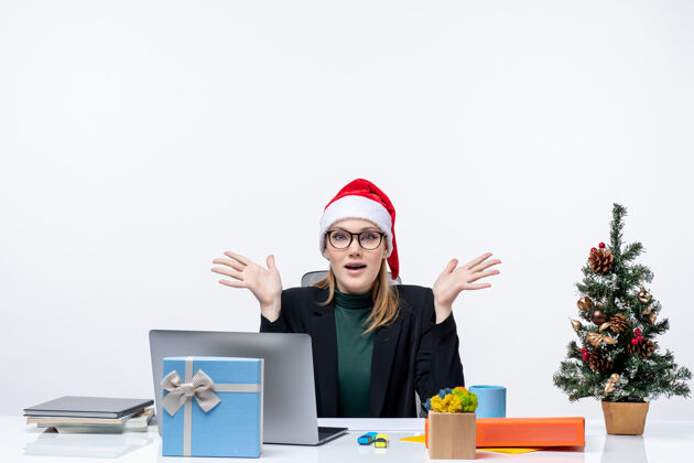 坐着新年心情坚定迷人的女人戴着圣诞老人帽坐在一张桌子旁 桌上放着一棵圣诞树和一份礼物在办公室里礼物迷人圣诞节