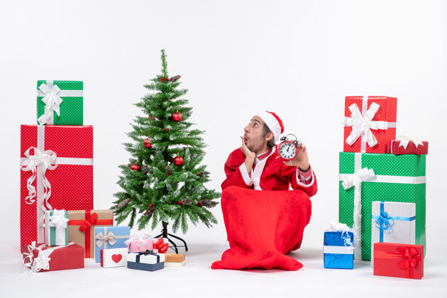 年轻人惊讶的年轻人坐在地上庆祝新年或圣诞节 拿着礼物和白色背景装饰圣诞树附近的时钟地圣诞树抱着